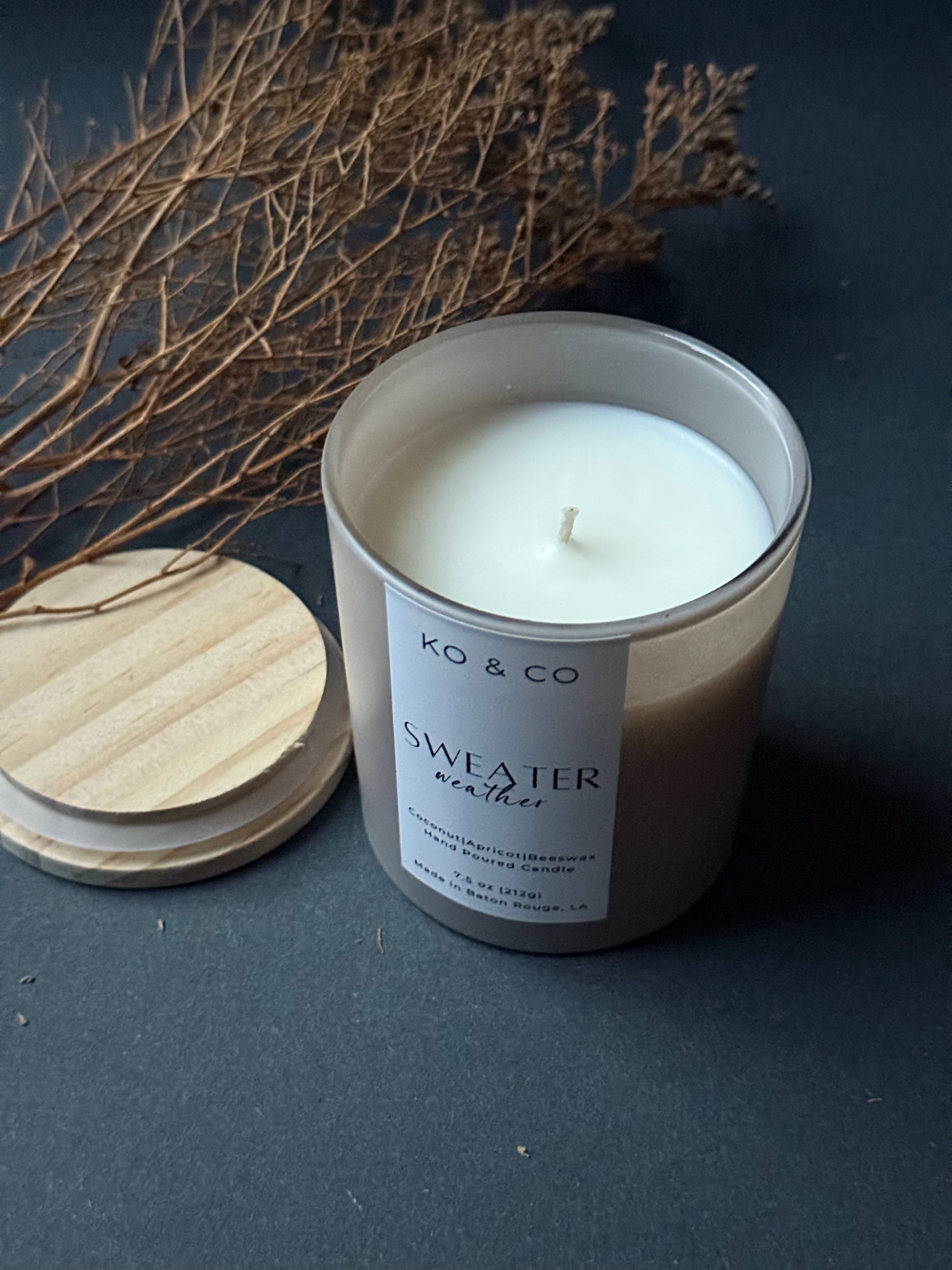 Candle with a label on a dark surface next to wooden lid and dried branches